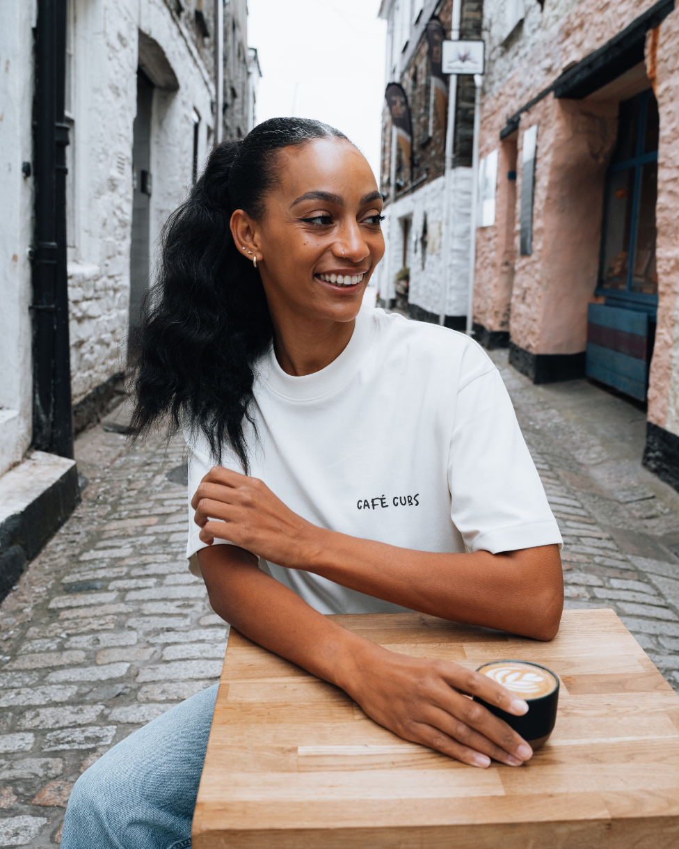 Woman sitting at a table with a coffee cup in an alleyway, wearing a &