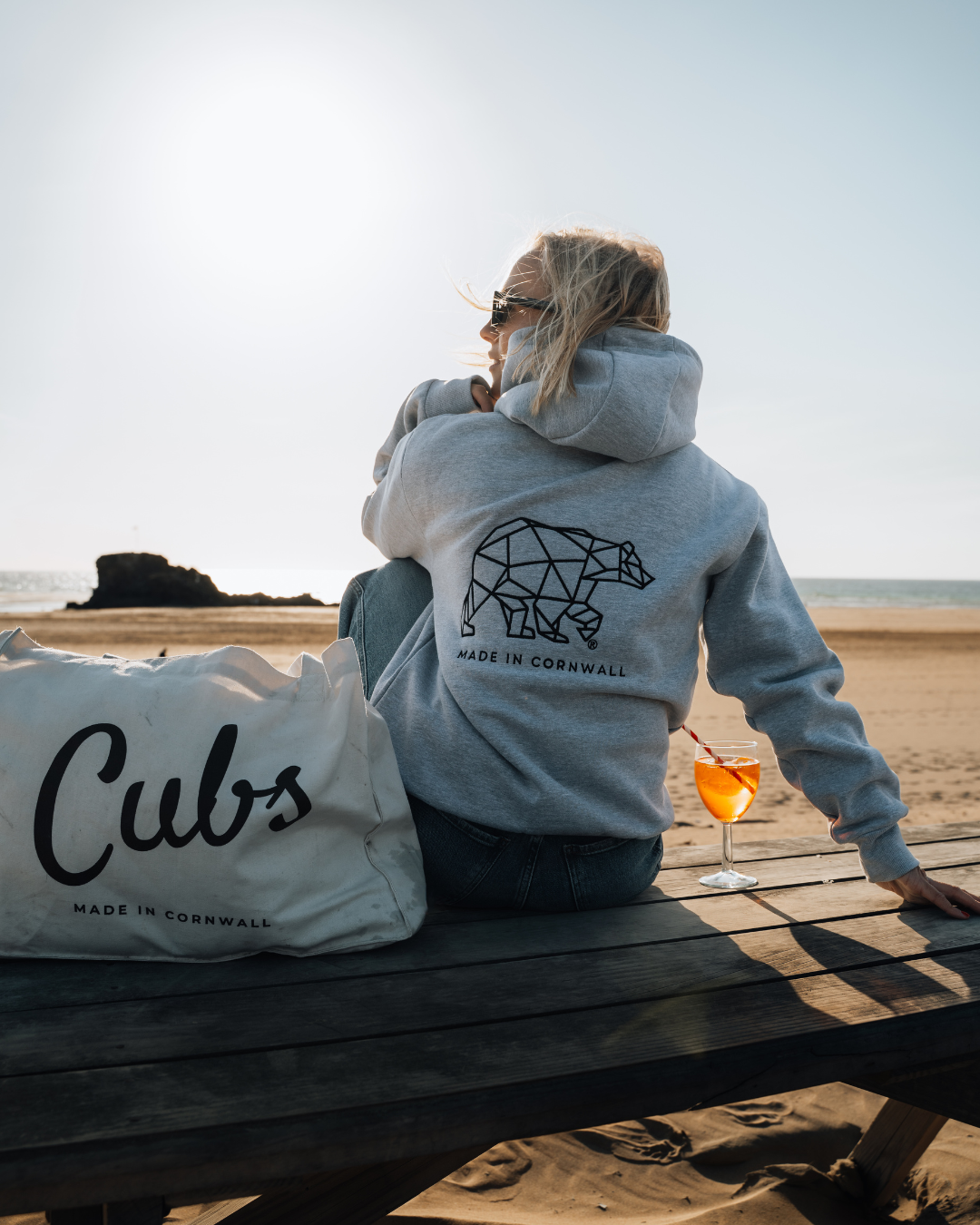 Person wearing a hoodie with a bear design on a beach, holding a drink and next to a &