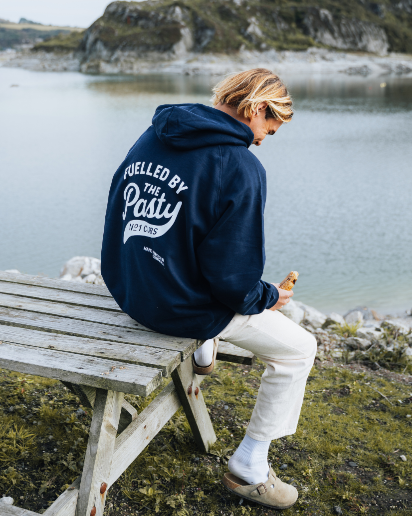 Person wearing a Cornish pasty hoodie with sitting on a bench by a lake