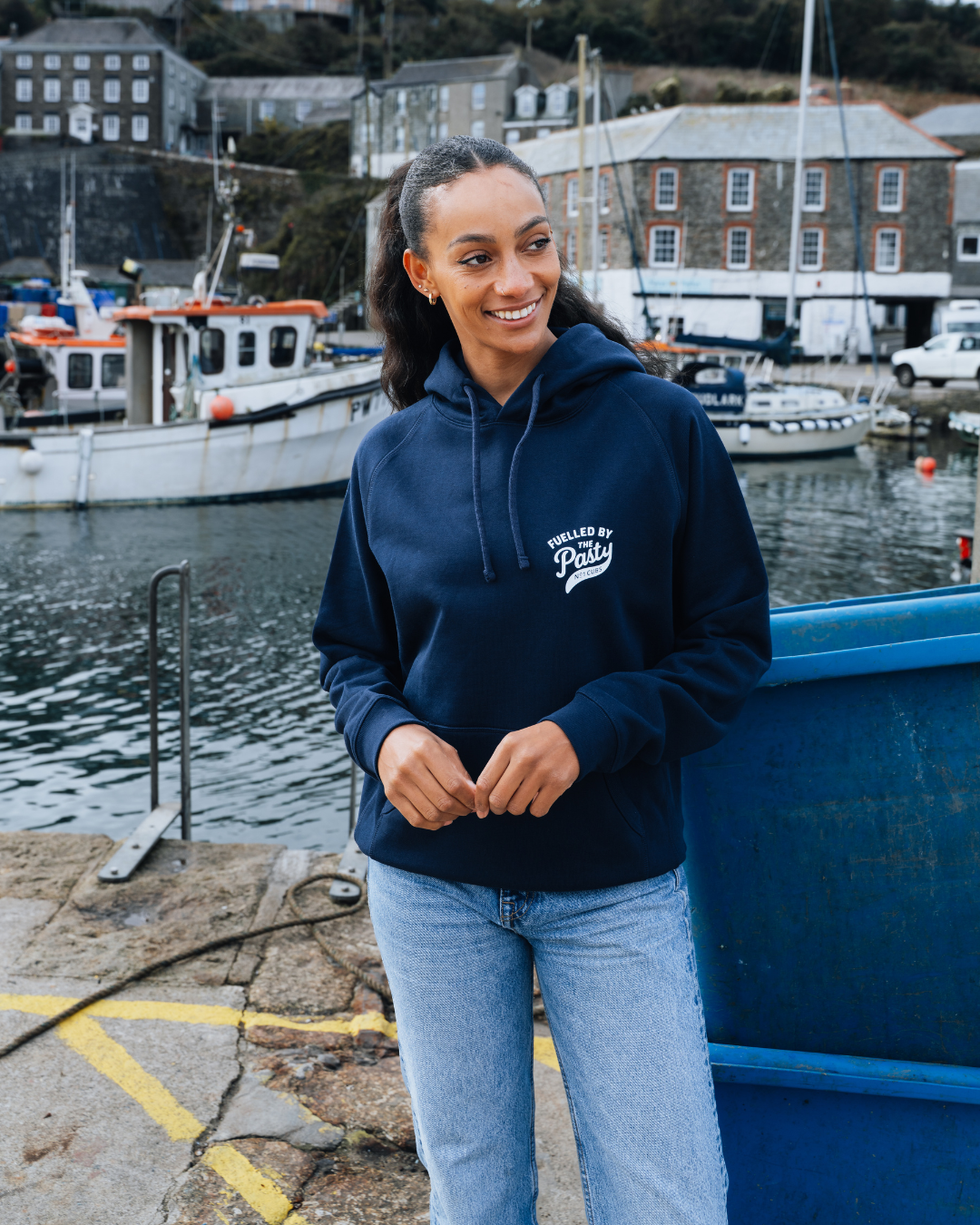 Woman wearing a navy hoodie with text, standing by a harbor with boats and buildings in the background.