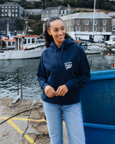 Woman wearing a navy hoodie with text, standing by a harbor with boats and buildings in the background.