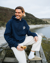 Man sitting on a wooden bench by a body of water, wearing a navy hoodie and white pants.